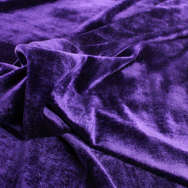Op de afbeelding: Een close-up van een dieppaarse fluwelen stof met een zachte, pluche textuur. De stof is gedrapeerd en gevouwen, wat een luxe en elegante uitstraling cre&euml;ert.