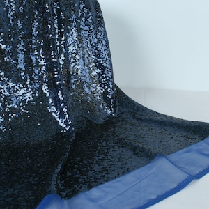 Pode incluir: Tecido de lantejoulas azul marinho com acabamento em malha azul. O tecido é drapeado, mostrando as lantejoulas brilhantes. O material é adequado para roupas ou projetos decorativos. As lantejoulas criam um efeito texturizado e luxuoso.