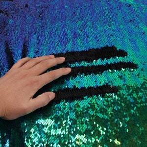 May include: A close-up of iridescent green and blue sequins on a fabric. The sequins are arranged in a pattern of rows, with a black stripe running horizontally across the fabric.