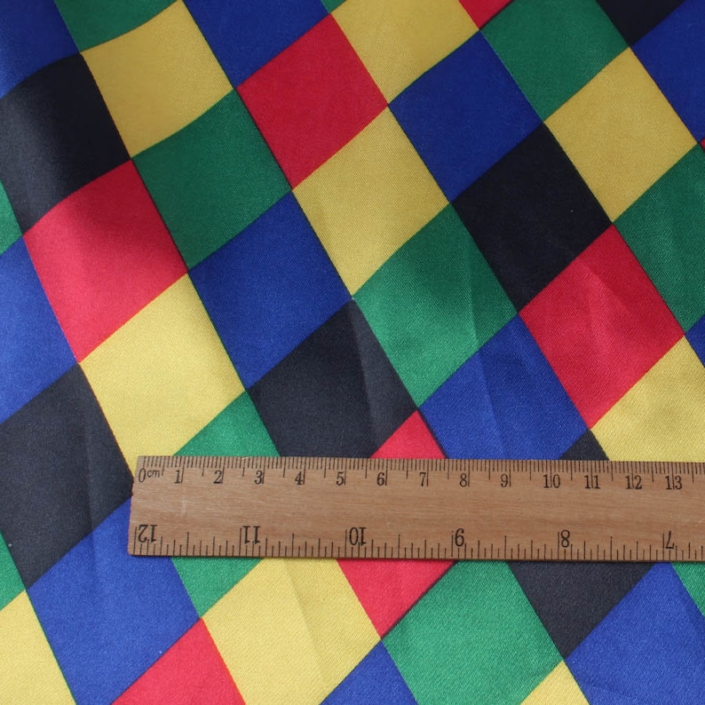 May include: A colorful fabric with a harlequin pattern of red, yellow, green, blue, and black diamond shapes. A wooden ruler with centimeter markings is placed across the fabric, showing measurements.