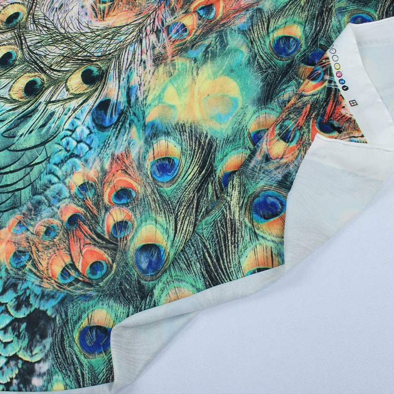 May include: Close-up of a fabric with a peacock feather print. The fabric is a light blue color with a white background. The peacock feathers are a vibrant green, blue, and orange.