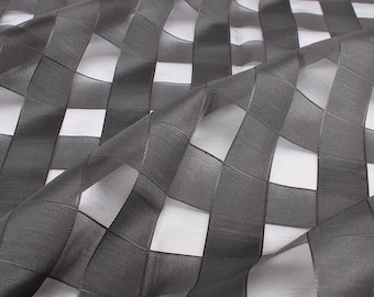May include: Close-up of a sheer fabric with a geometric checkered pattern. The fabric is a dark gray color, with a grid of squares and rectangles. The fabric is folded, creating shadows and highlights.