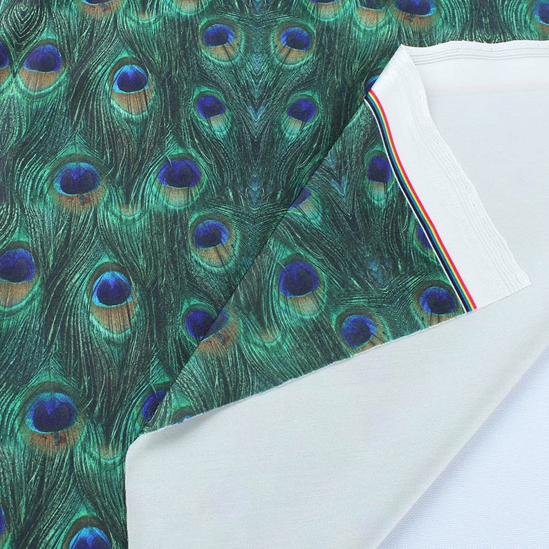 May include: Close-up of a fabric with a peacock feather print. The fabric is green with blue and brown accents. The fabric is folded over to show the white underside.