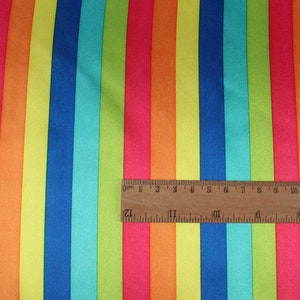 May include: Vibrant fabric with vertical stripes in rainbow colors: orange, yellow, green, blue, and pink. A wooden ruler is placed horizontally across the fabric, showing centimeter measurements.