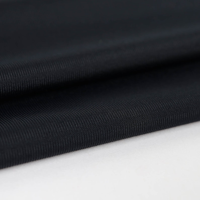 May include: Close-up of a black, textured fabric with a subtle ribbed pattern. The fabric is folded and draped, showcasing its smooth and soft texture.