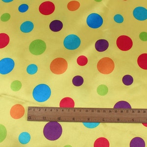 May include: Yellow fabric with a pattern of colorful polka dots in red, orange, blue, green, and purple. A wooden ruler is placed across the fabric, showing measurements in inches.