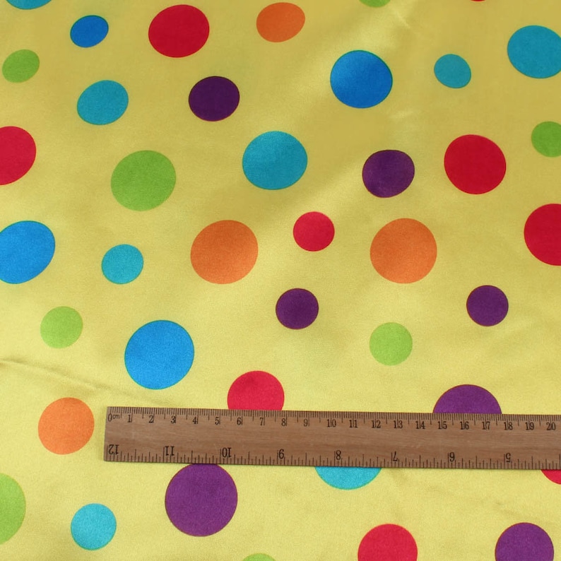 May include: Yellow fabric with a pattern of colorful polka dots in red, orange, blue, green, and purple. A wooden ruler is placed across the fabric, showing measurements in inches.
