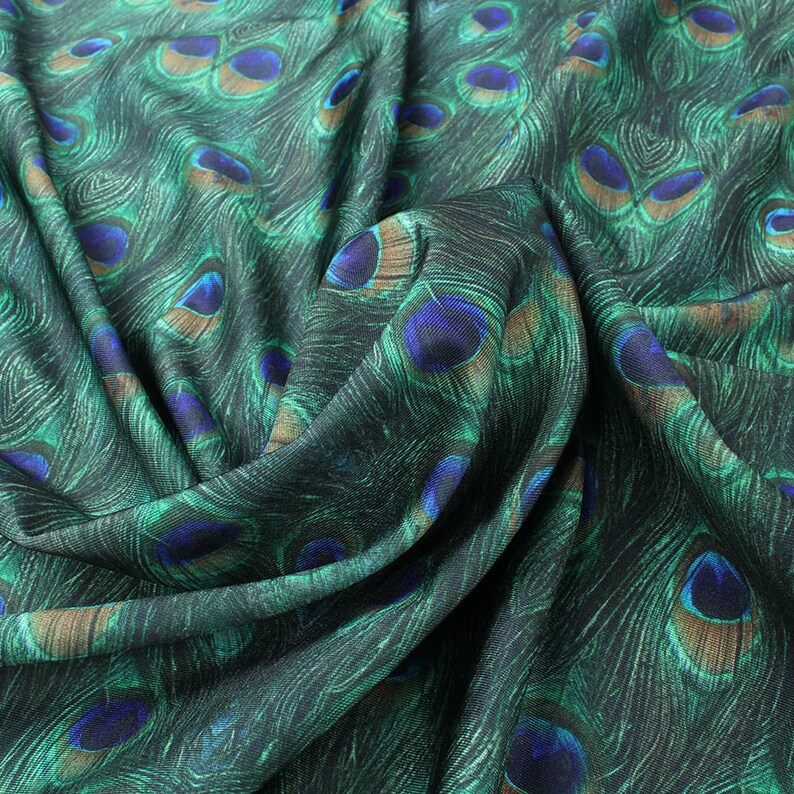 May include: Green fabric with a peacock feather pattern. The fabric is folded and draped, showing the texture and detail of the print.