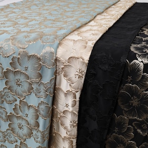 May include: Three folded lengths of fabric featuring a raised floral pattern. The first is light blue with gold flowers, the second is cream with gold flowers, and the third is black with gold flowers. Each piece displays a repeating botanical design.