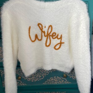 Puede incluir: Un su&eacute;ter crema y esponjoso con mangas largas y corte corto. La palabra "Wifey" est&aacute; bordada en hilo dorado en el pecho. El su&eacute;ter est&aacute; colgado de una percha de madera.