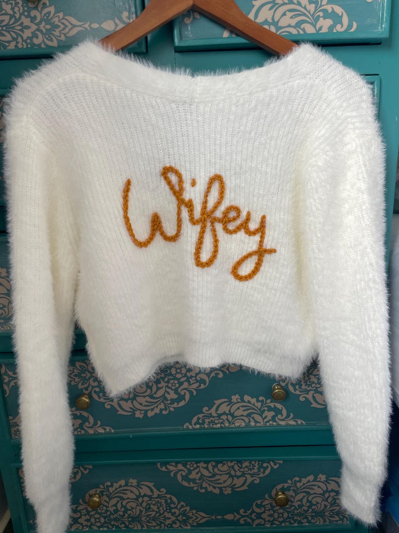 Puede incluir: Un su&eacute;ter crema y esponjoso con mangas largas y corte corto. La palabra "Wifey" est&aacute; bordada en hilo dorado en el pecho. El su&eacute;ter est&aacute; colgado de una percha de madera.
