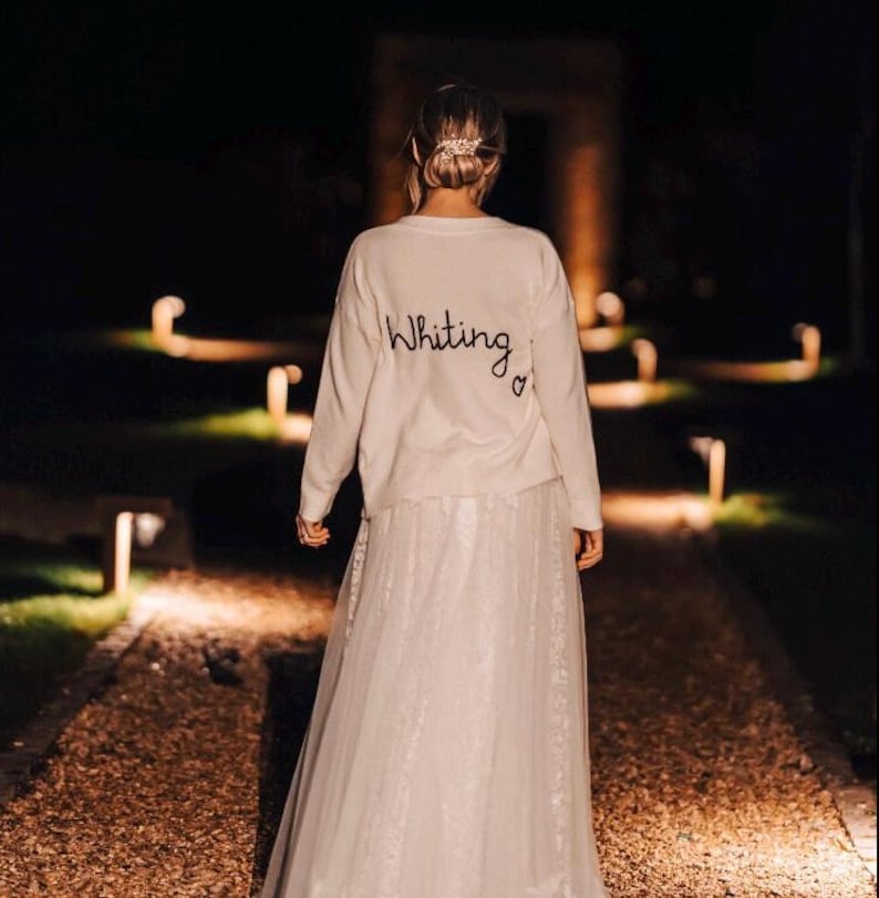 May include: A white sweater with the text "Whiting" and a heart embroidered on the back. The sweater is worn by a person with their back to the camera, walking on a path lit by lights.
