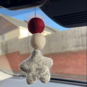 May include: A grey felt star with a white felt ball and a burgundy felt ball hanging from it. The star is hanging from a rear view mirror in a car.