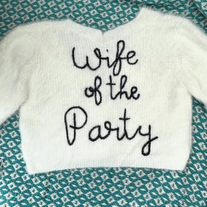 Puede incluir: Un su&eacute;ter blanco y esponjoso con mangas largas. La frase "Wife of the Party" est&aacute; bordada en negro en la parte delantera. El su&eacute;ter se muestra sobre una superficie estampada de color turquesa.