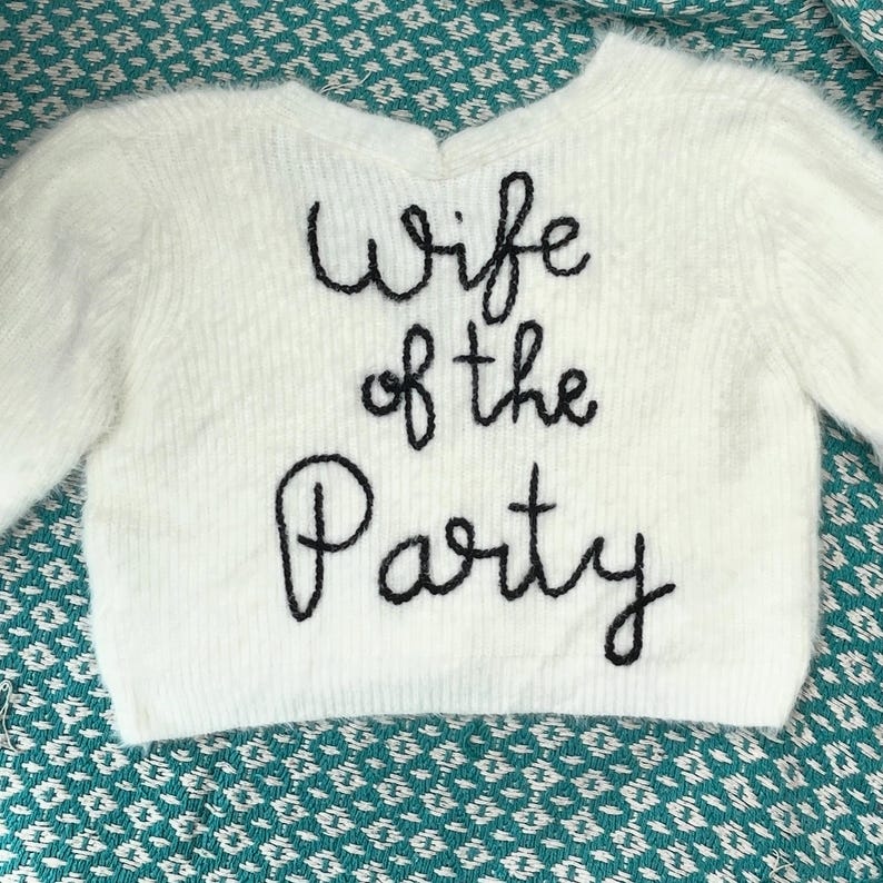 Puede incluir: Un su&eacute;ter blanco y esponjoso con mangas largas. La frase "Wife of the Party" est&aacute; bordada en negro en la parte delantera. El su&eacute;ter se muestra sobre una superficie estampada de color turquesa.