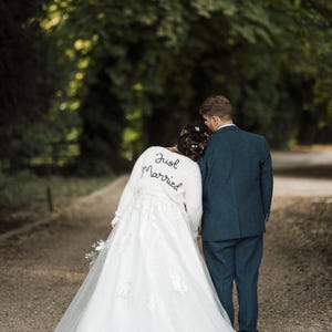 Puede incluir: Una pareja de reci&eacute;n casados camina por un sendero, vista desde atr&aacute;s. La novia lleva un vestido de novia blanco con detalles de encaje y una chaqueta blanca y esponjosa con "Just Married" bordado. El novio viste un traje azul oscuro.