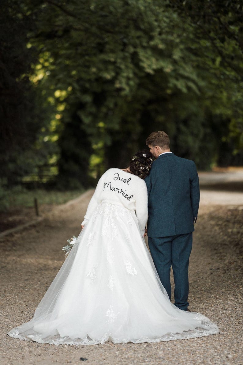 Puede incluir: Una pareja de reci&eacute;n casados camina por un sendero, vista desde atr&aacute;s. La novia lleva un vestido de novia blanco con detalles de encaje y una chaqueta blanca y esponjosa con "Just Married" bordado. El novio viste un traje azul oscuro.