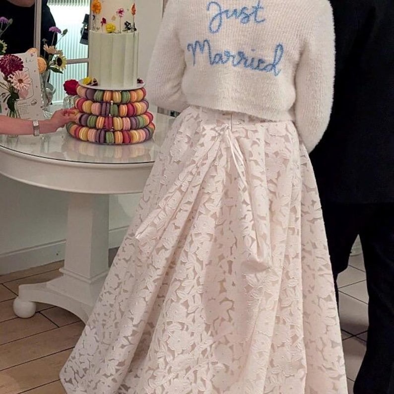 Puede incluir: Una novia con una falda de encaje blanco y un su&eacute;ter blanco esponjoso con "Just Married" bordado en azul. Una tarta de varios pisos con flores y macarons est&aacute; sobre una mesa blanca.