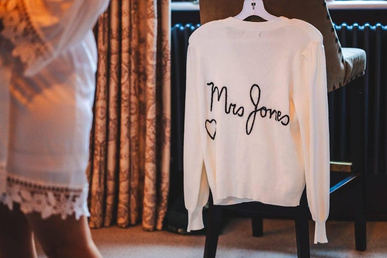 Puede incluir: Su&eacute;ter color crema con mangas largas, colgado de una percha. La parte trasera del su&eacute;ter presenta las palabras bordadas "Mrs Jones" en escritura negra, con un peque&ntilde;o coraz&oacute;n. El su&eacute;ter se exhibe en una silla negra.