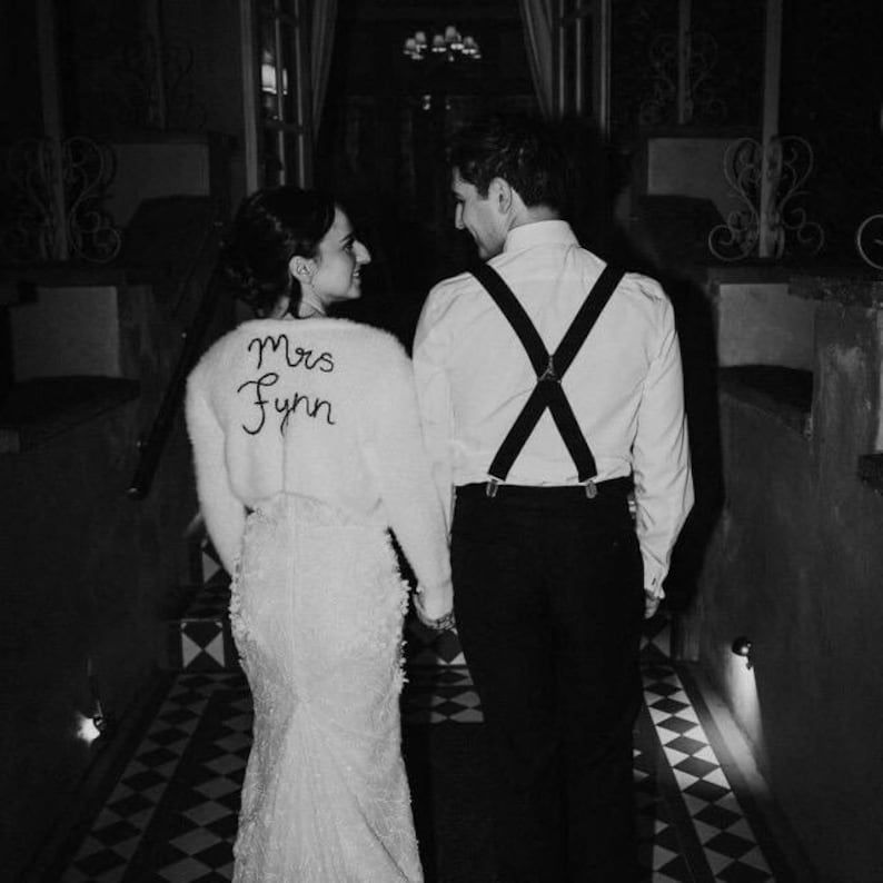 Puede incluir: Imagen en blanco y negro de una pareja caminando por un pasillo. La mujer lleva un vestido blanco y un su&eacute;ter blanco con "Mrs Fynn" bordado en la espalda. El hombre lleva camisa blanca, pantal&oacute;n negro y tirantes.