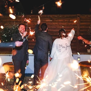 Puede incluir: Celebraci&oacute;n nocturna con bengalas. Una persona lleva una chaqueta blanca con "Mrs. Jones" y un coraz&oacute;n en la espalda. La escena est&aacute; iluminada por las chispas y la luz c&aacute;lida, creando un ambiente festivo.
