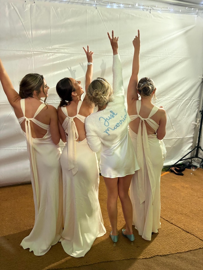 Puede incluir: Cinco personas celebrando, vestidas con vestidos de color claro y una bata corta. La bata tiene las palabras "Just Married" bordadas en la espalda. Los vestidos tienen la espalda baja con un detalle de lazo. Las personas levantan los brazos en se&ntilde;al de celebraci&oacute;n.