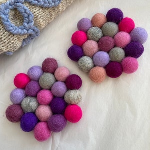 May include: Two round coasters made of felt balls in shades of pink, purple, and grey. The balls are arranged in a circular pattern.