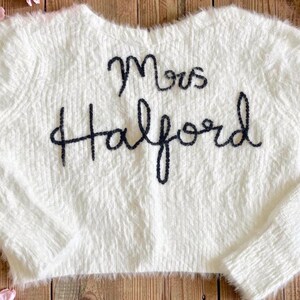 Puede incluir: Un su&eacute;ter blanco y esponjoso con el texto bordado "Mrs. Halford" en azul oscuro. El su&eacute;ter se muestra sobre una superficie de madera r&uacute;stica, rodeado de flores rosas y vegetaci&oacute;n.