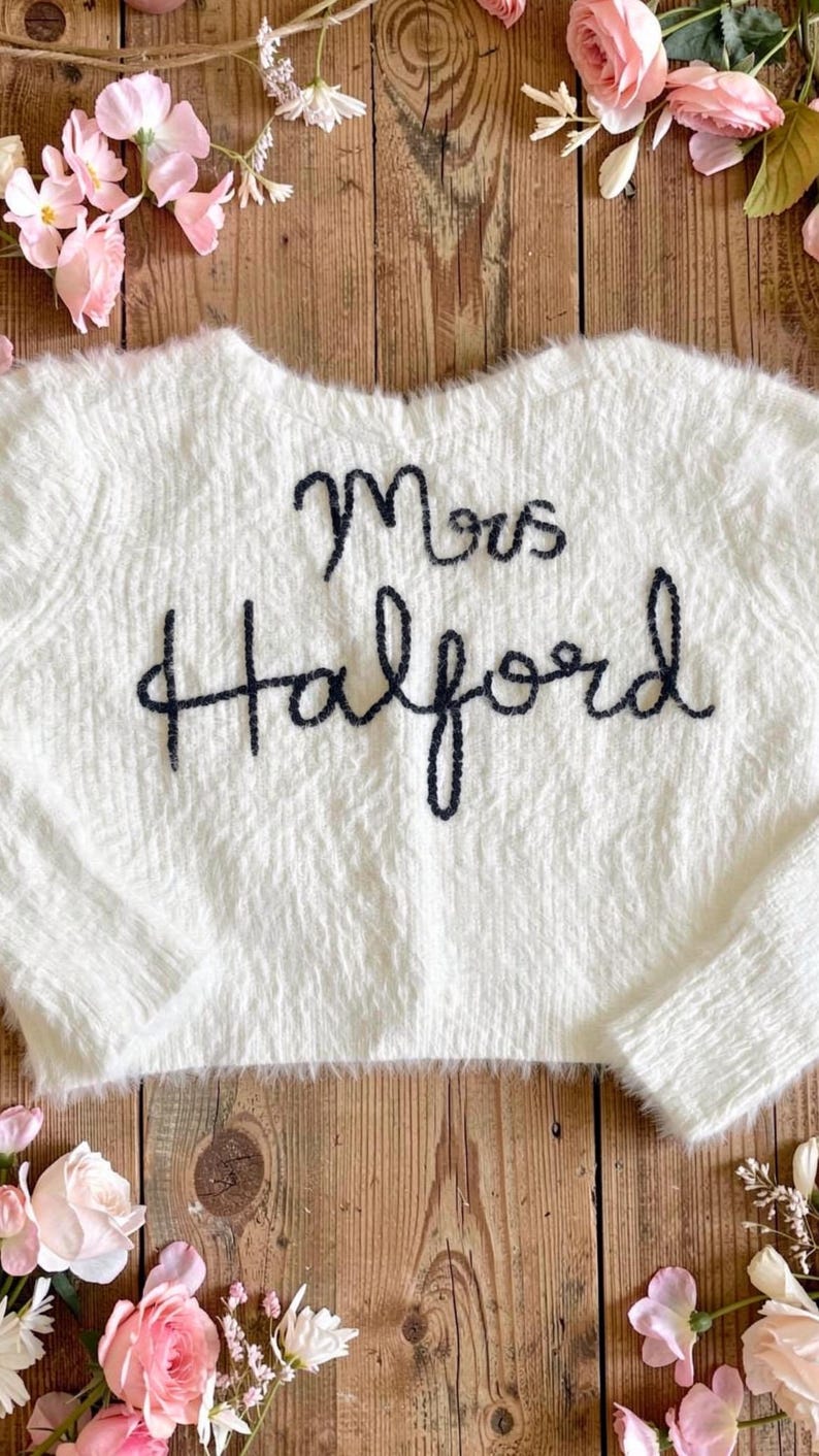 Puede incluir: Un su&eacute;ter blanco y esponjoso con el texto bordado "Mrs. Halford" en azul oscuro. El su&eacute;ter se muestra sobre una superficie de madera r&uacute;stica, rodeado de flores rosas y vegetaci&oacute;n.