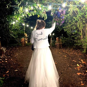Puede incluir: Una novia con un vestido de novia blanco y una chaqueta blanca con "Just Married" bordado en la espalda, sosteniendo un ramo de flores. La escena es al aire libre, con luces de cadena y exuberante vegetaci&oacute;n en el fondo.