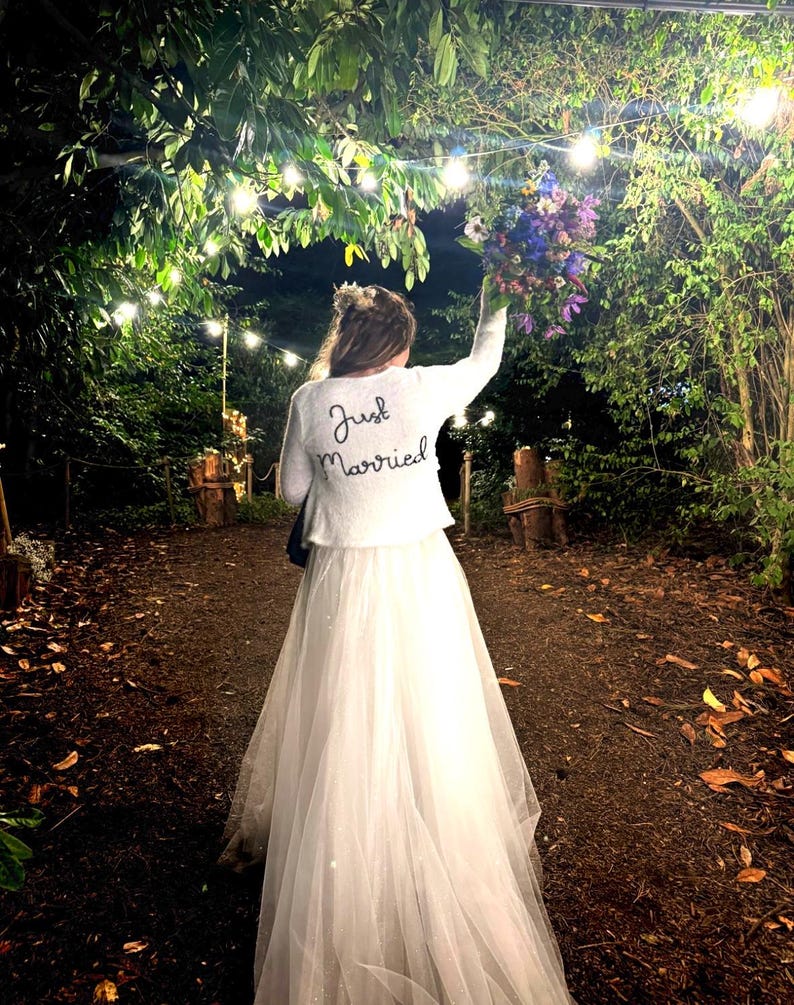 Puede incluir: Una novia con un vestido de novia blanco y una chaqueta blanca con "Just Married" bordado en la espalda, sosteniendo un ramo de flores. La escena es al aire libre, con luces de cadena y exuberante vegetaci&oacute;n en el fondo.
