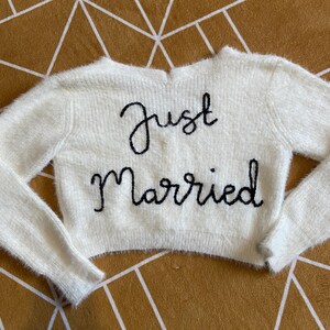 Puede incluir: Un su&eacute;ter crema y difuso con mangas largas. Las palabras "Just Married" est&aacute;n bordadas en negro en la parte delantera. El su&eacute;ter se muestra sobre una superficie con un patr&oacute;n geom&eacute;trico dorado y blanco.