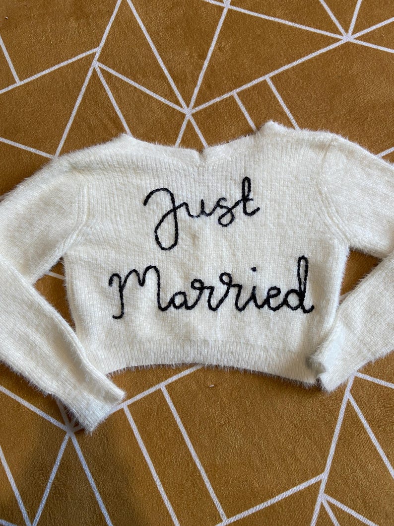 Puede incluir: Un su&eacute;ter crema y difuso con mangas largas. Las palabras "Just Married" est&aacute;n bordadas en negro en la parte delantera. El su&eacute;ter se muestra sobre una superficie con un patr&oacute;n geom&eacute;trico dorado y blanco.