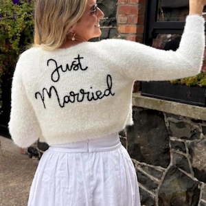 Puede incluir: Un su&eacute;ter corto blanco y esponjoso con las palabras "Just Married" bordadas en escritura negra. El su&eacute;ter se combina con una falda blanca. La imagen sugiere un tema de boda o novia, capturando un momento de celebraci&oacute;n.