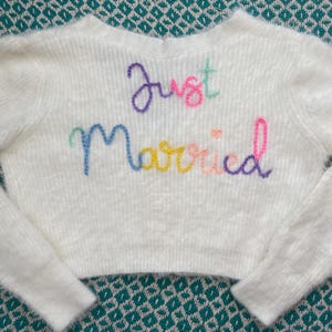 Puede incluir: Un su&eacute;ter blanco corto con mangas largas. Las palabras "Just Married" est&aacute;n bordadas en la parte delantera con hilo colorido. El su&eacute;ter est&aacute; hecho de un material suave y texturizado, ideal para una boda o evento nupcial.