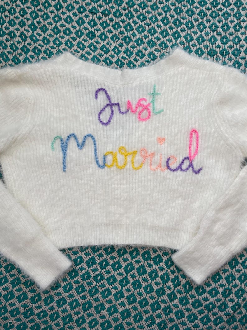 Puede incluir: Un su&eacute;ter blanco corto con mangas largas. Las palabras "Just Married" est&aacute;n bordadas en la parte delantera con hilo colorido. El su&eacute;ter est&aacute; hecho de un material suave y texturizado, ideal para una boda o evento nupcial.