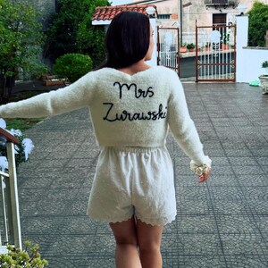 Puede incluir: Un su&eacute;ter crema difuso con el nombre "Mrs. Zurawski" bordado en escritura negra en la espalda. El su&eacute;ter se combina con pantalones cortos blancos. La persona est&aacute; de pie en una escalera de piedra.