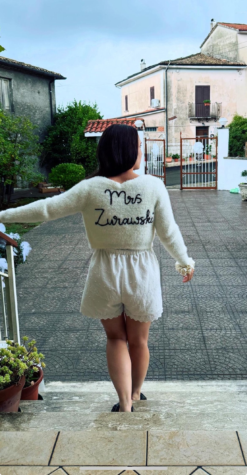 Puede incluir: Un su&eacute;ter crema difuso con el nombre "Mrs. Zurawski" bordado en escritura negra en la espalda. El su&eacute;ter se combina con pantalones cortos blancos. La persona est&aacute; de pie en una escalera de piedra.