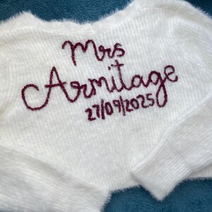 Puede incluir: Un su&eacute;ter corto blanco y esponjoso con el texto bordado "Mrs Armitage" y la fecha "27/09/2025" en color burdeos. El su&eacute;ter tiene mangas largas y una textura suave.