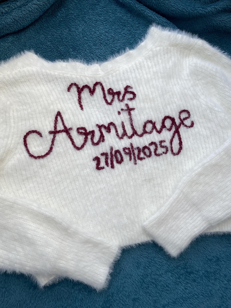Puede incluir: Un su&eacute;ter corto blanco y esponjoso con el texto bordado "Mrs Armitage" y la fecha "27/09/2025" en color burdeos. El su&eacute;ter tiene mangas largas y una textura suave.
