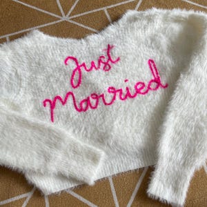 Puede incluir: Un su&eacute;ter blanco y esponjoso con las palabras "Just Married" bordadas en rosa brillante en el pecho. El su&eacute;ter tiene mangas largas y un corte corto, perfecto para una reci&eacute;n casada.