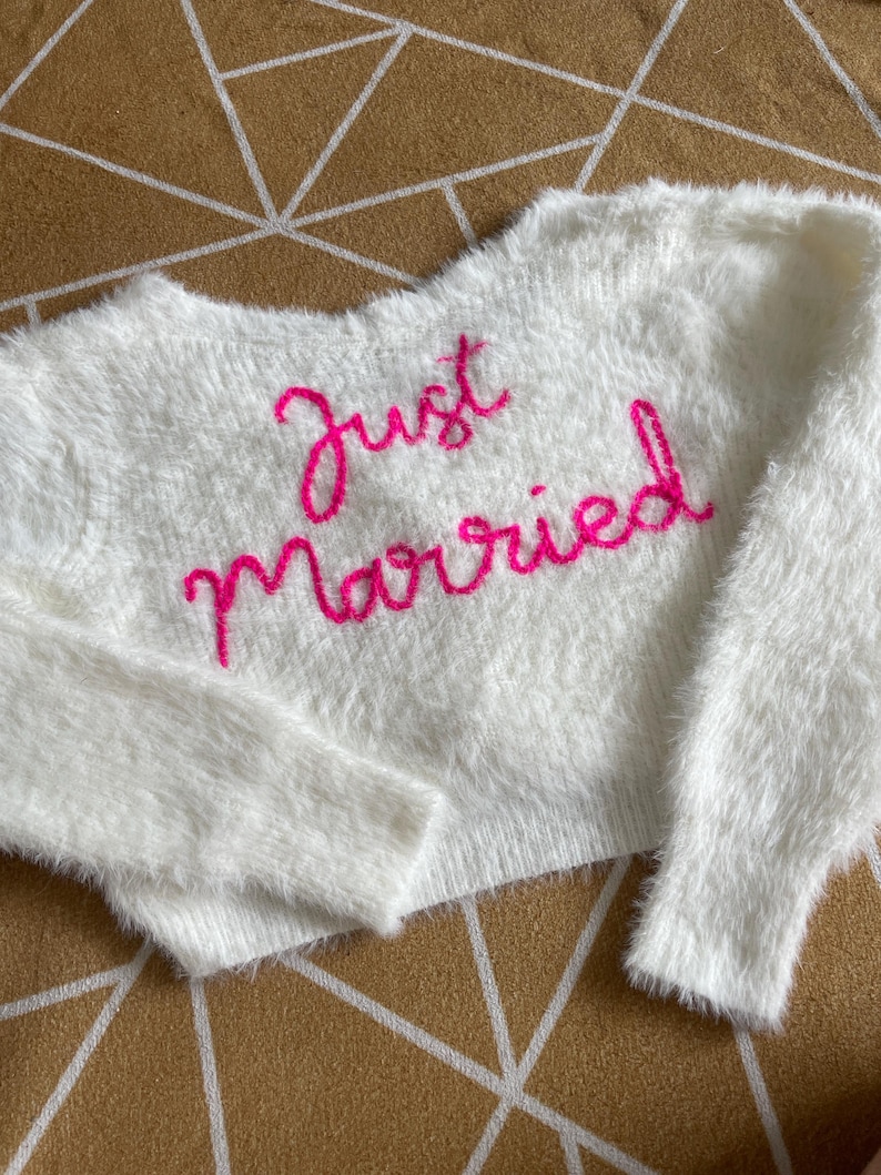 Puede incluir: Un su&eacute;ter blanco y esponjoso con las palabras "Just Married" bordadas en rosa brillante en el pecho. El su&eacute;ter tiene mangas largas y un corte corto, perfecto para una reci&eacute;n casada.