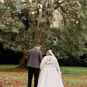 May include: A bride and groom walking away from the camera in a park. The bride is wearing a white wedding dress with a veil and a jacket that says "Just Married" on the back. The groom is wearing a gray suit jacket and black pants.
