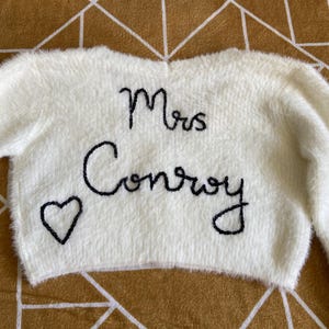 Puede incluir: Un su&eacute;ter esponjoso de color crema con mangas largas. Las palabras "Mrs Conroy" est&aacute;n bordadas en negro, junto con un peque&ntilde;o coraz&oacute;n. Es una prenda de tem&aacute;tica nupcial o de boda.