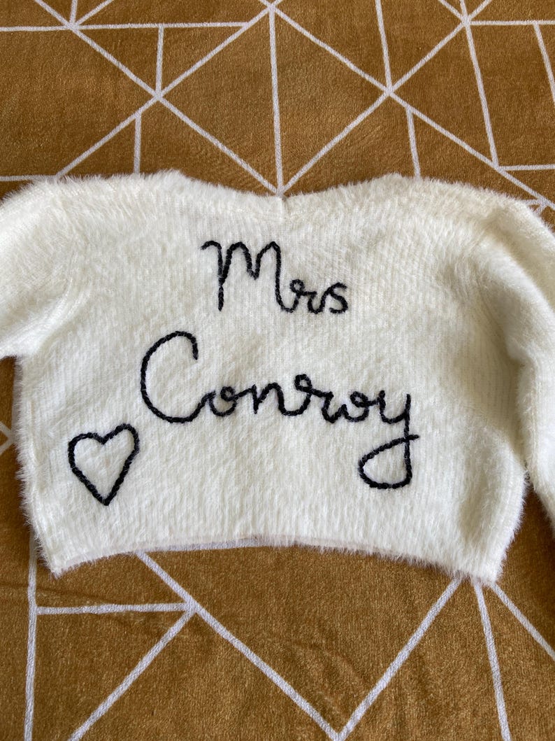 Puede incluir: Un su&eacute;ter esponjoso de color crema con mangas largas. Las palabras "Mrs Conroy" est&aacute;n bordadas en negro, junto con un peque&ntilde;o coraz&oacute;n. Es una prenda de tem&aacute;tica nupcial o de boda.