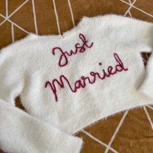 Puede incluir: Un su&eacute;ter blanco y esponjoso con las palabras "Just Married" bordadas en un guion rosa intenso. El su&eacute;ter tiene mangas largas y un corte corto. El fondo es una superficie marr&oacute;n con un patr&oacute;n geom&eacute;trico.