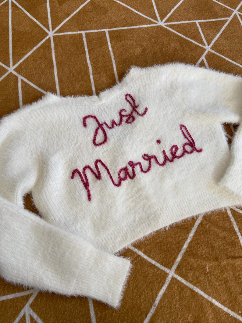 Puede incluir: Un su&eacute;ter blanco y esponjoso con las palabras "Just Married" bordadas en un guion rosa intenso. El su&eacute;ter tiene mangas largas y un corte corto. El fondo es una superficie marr&oacute;n con un patr&oacute;n geom&eacute;trico.