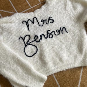 Puede incluir: Un su&eacute;ter blanco y esponjoso con el texto bordado "Mrs Benson" en cursiva negra. El su&eacute;ter tiene cuello redondo y mangas largas. La tela parece suave y texturizada.
