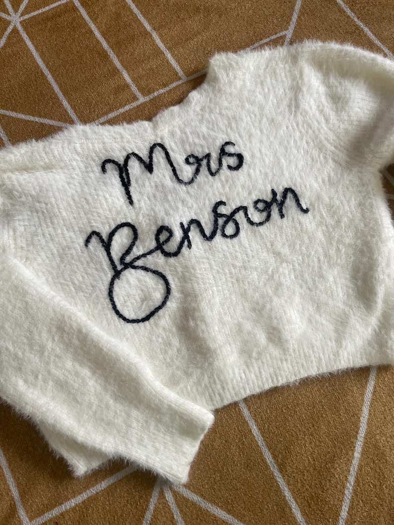 Puede incluir: Un su&eacute;ter blanco y esponjoso con el texto bordado "Mrs Benson" en cursiva negra. El su&eacute;ter tiene cuello redondo y mangas largas. La tela parece suave y texturizada.