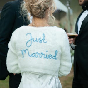 Puede incluir: Un bolero blanco y esponjoso con las palabras "Just Married" bordadas en azul claro. El su&eacute;ter se usa sobre un vestido blanco. La imagen est&aacute; tomada en una boda al aire libre.